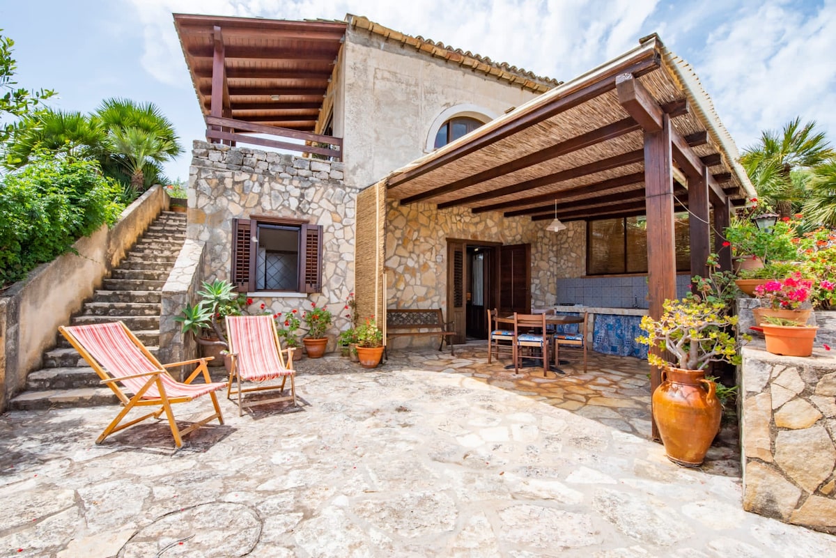 An outdoor patio area is presented with stone flooring, surrounded by lush greenery and colorful potted plants. Two striped lounge chairs are positioned for relaxation. A dining table with chairs is visible, under a shaded terrace, providing an inviting space for enjoying meals outdoors.