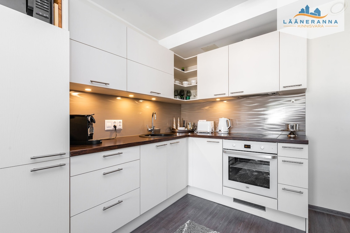 A modern kitchen is presented with sleek white cabinets and a dark countertop. Under-cabinet lighting enhances the workspace, which includes essential appliances like a coffee maker and an oven. Open shelving displays neatly arranged dishes, contributing to a tidy appearance.