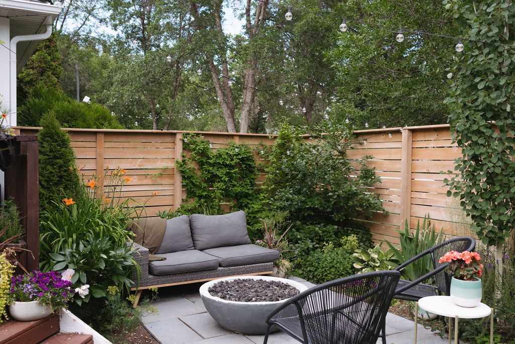 A serene garden oasis is displayed with a gray sofa surrounded by vibrant greenery and blooming flowers. A circular stone fire pit is positioned at the center, complemented by two stylish black chairs. String lights hang overhead, adding a cozy ambiance to the outdoor space.