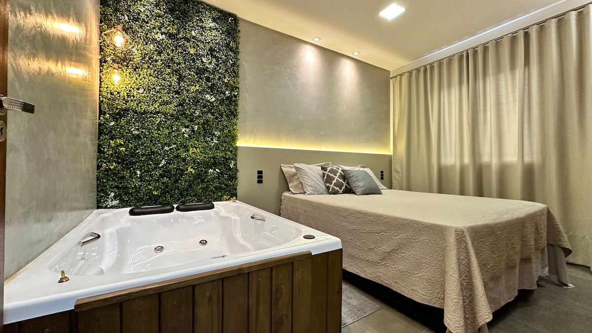 A spacious sleeping area is complemented by a cozy bed, adorned with neutral bedding. Adjacent, a private hot tub features wood paneling and is positioned against a green accent wall. Soft lighting enhances the relaxing ambiance, with sheer curtains allowing natural light to filter in.