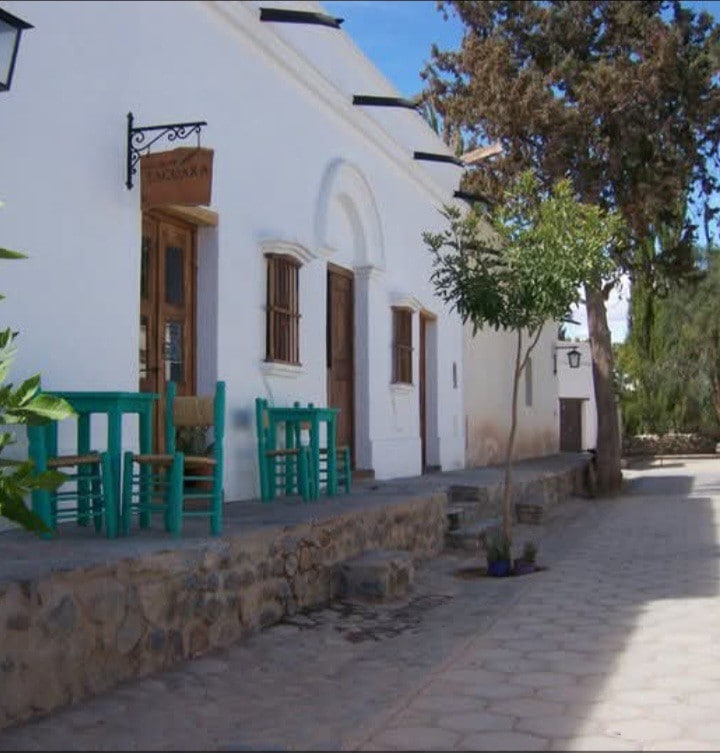 A charming white building with wooden doors and windows is depicted alongside a cobblestone pathway. Colorful turquoise tables and chairs are positioned on a stone platform, surrounded by greenery and shaded by trees, creating a welcoming outdoor space.