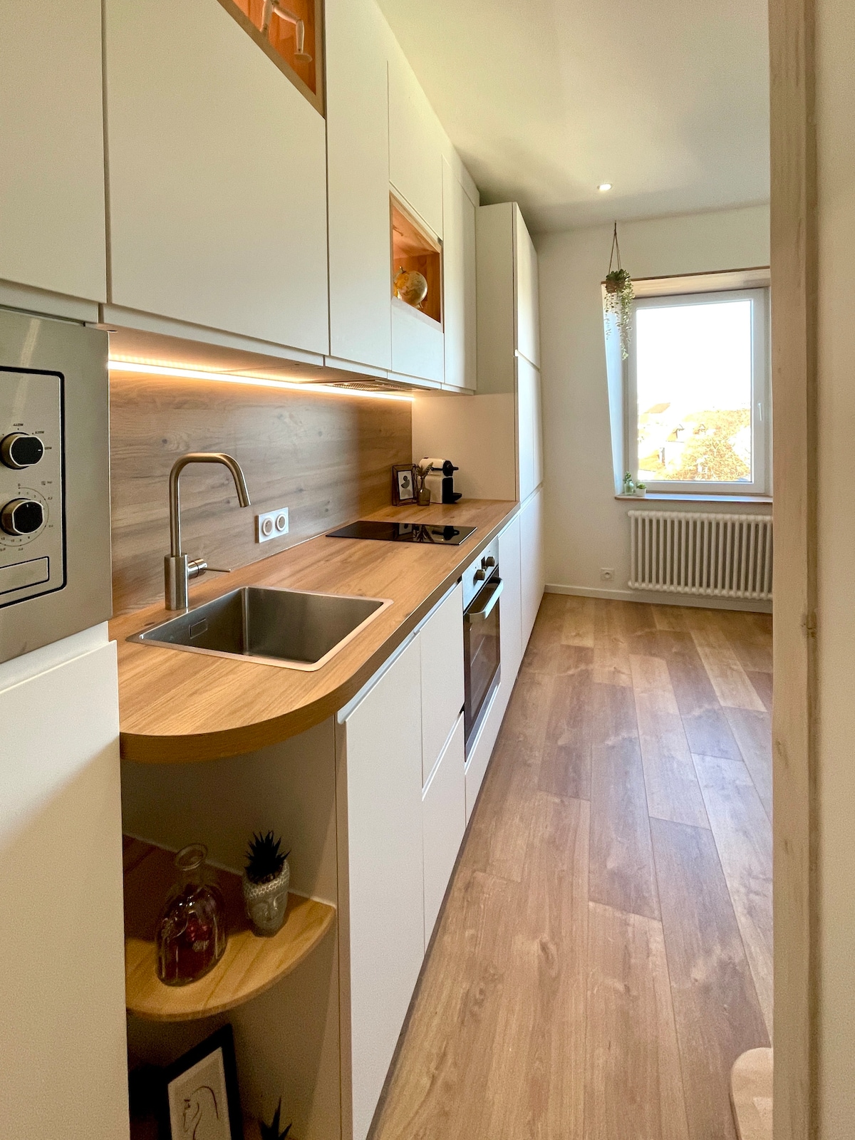 A compact kitchen features modern cabinetry with wooden accents and ample counter space. Equipped with a stainless steel sink, built-in oven, and stovetop, it is bathed in natural light from the nearby window, enhancing its functional design.