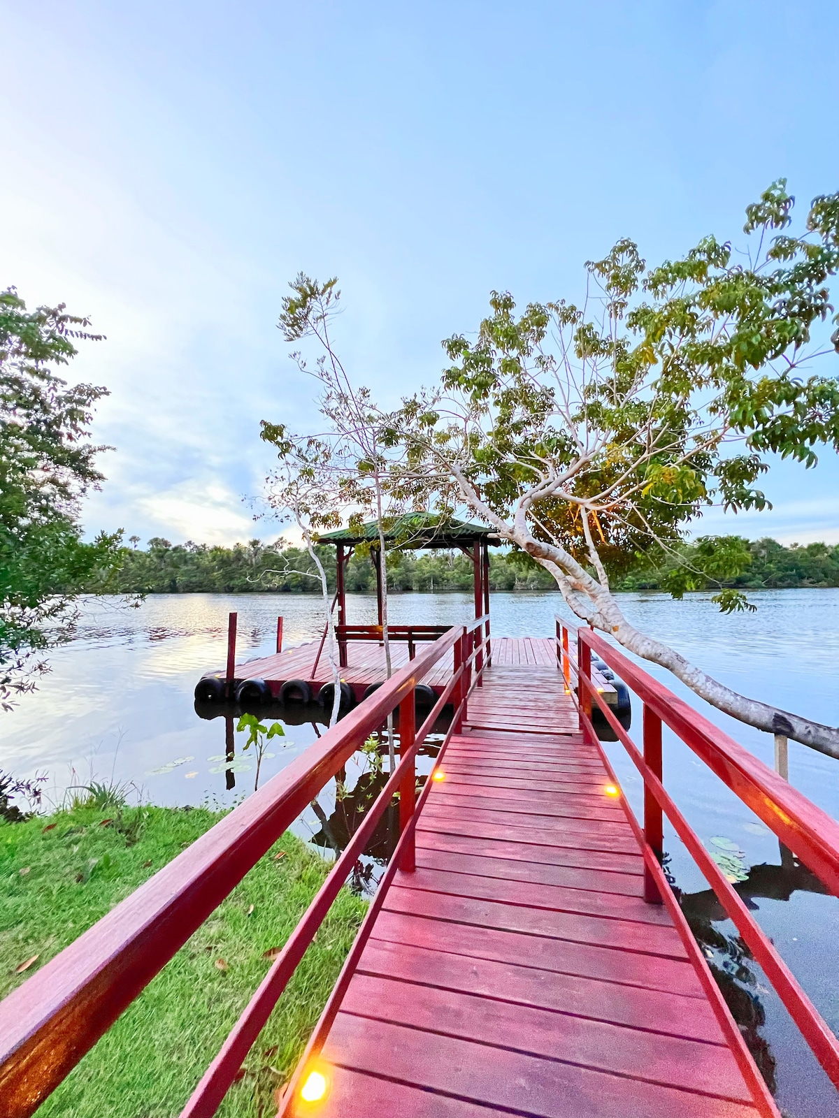 A wooden pier in vibrant red stretches over calm waters, surrounded by lush greenery. Soft lighting highlights the walkway, and a shaded area with support for boats is visible at the end. The tranquil setting invites relaxation by the river.