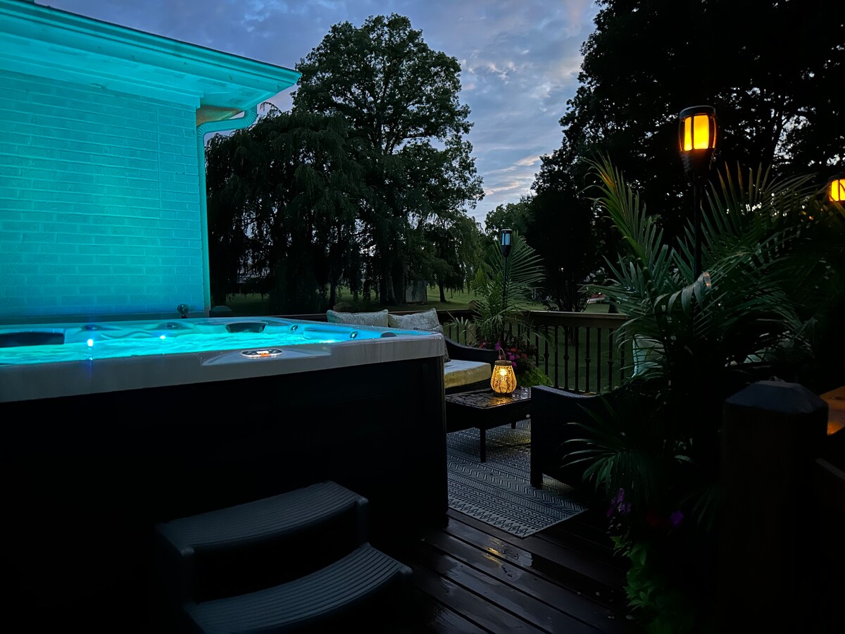 An outdoor space features a hot tub emitting a soft blue glow, surrounded by lush greenery. A cozy seating area is present nearby, with decorative lanterns providing gentle lighting. The deck, composed of warm wood, blends seamlessly into the serene landscape.