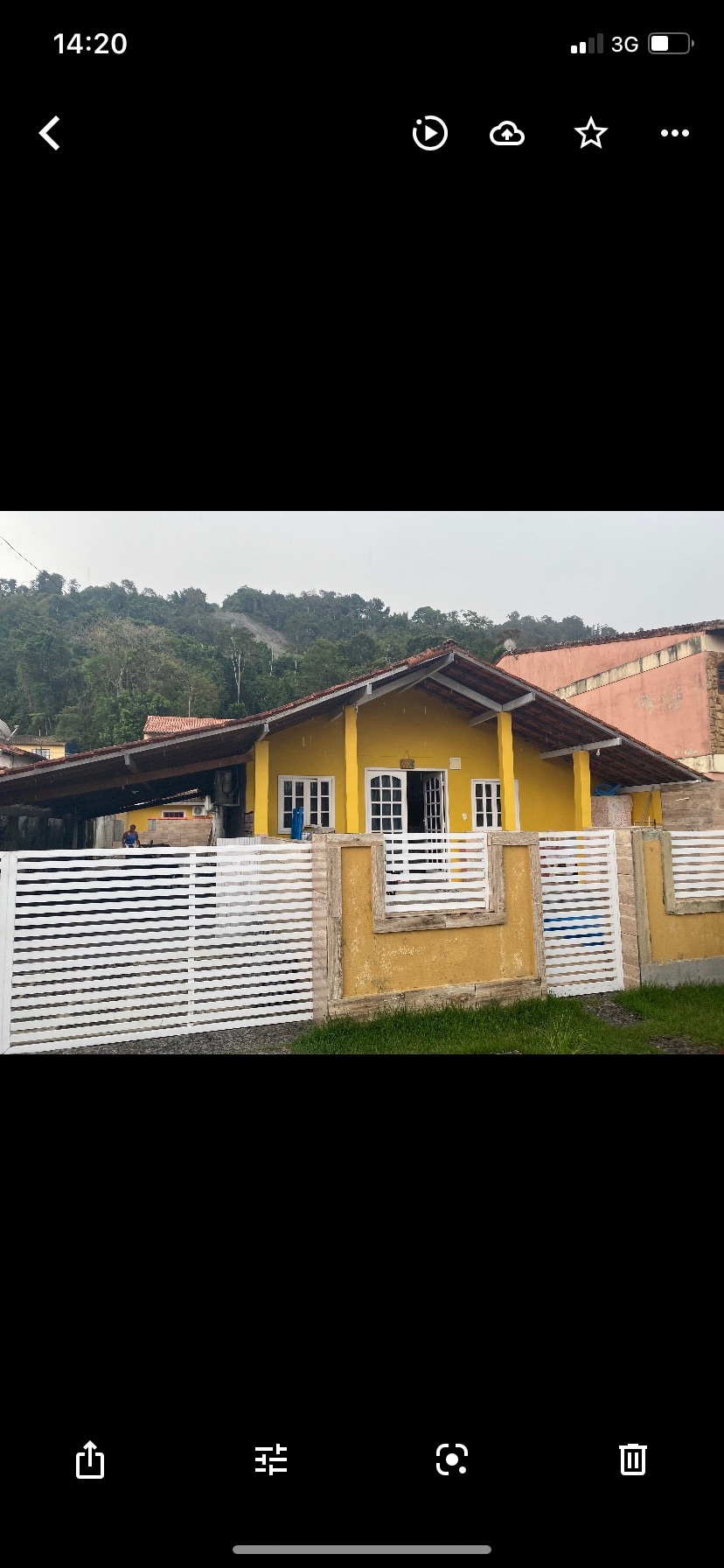 The exterior of a charming house is displayed, featuring a bright yellow façade. White wooden fencing surrounds the property, and the entrance is highlighted by large glass doors and windows. Lush greenery is visible in the background, creating a welcoming environment.