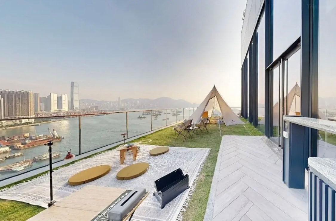 An expansive terrace overlooks a stunning view of the water and city skyline. Various seating options, including round tables and lounge chairs, are placed on a grassy area alongside an outdoor rug. A tent adds a cozy touch to the open-air space.