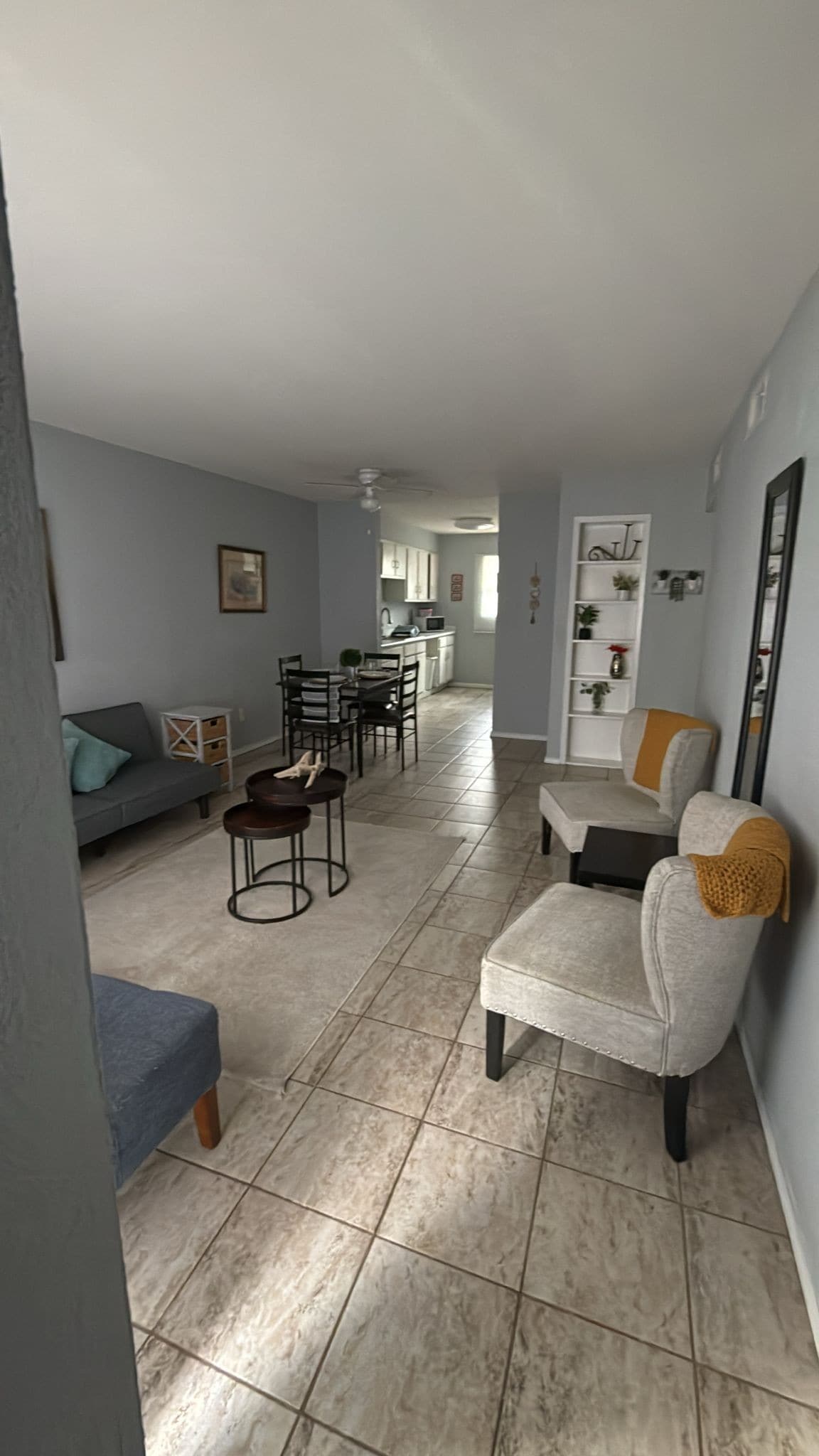 An open living space is seen, featuring a blend of seating options, including a gray futon and two chairs with yellow accents. A dining area with black chairs is in view, along with a kitchen that showcases light-colored cabinetry. Natural light illuminates the room.
