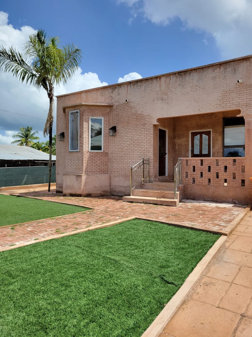The exterior of a single-story building is depicted, featuring a textured facade and multiple large windows. A small set of steps leads to the entrance. The surrounding area displays artificial grass and paved pathways, complemented by tropical palm trees in the background.