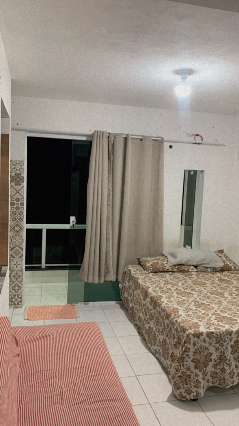 A cozy space is shown, featuring a spacious bed dressed in a floral-patterned coverlet. A red-striped sofa sits nearby. Light-colored curtains frame a large window, allowing for natural light, while the tiled floor extends throughout the area, enhancing the sense of openness.