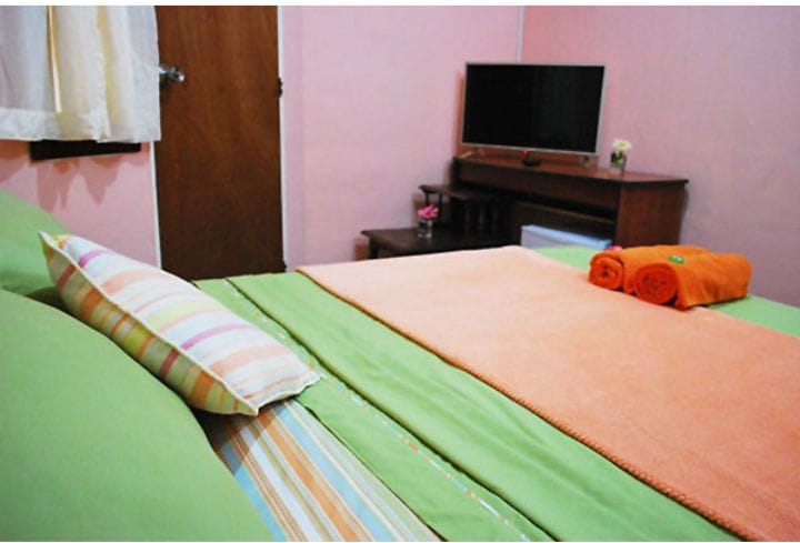 A cozy sleeping area features pastel pink walls, with a spacious bed dressed in vibrant green and orange linens. A striped pillow adds a touch of color. A wooden table with a television is visible, creating a functional and inviting atmosphere.