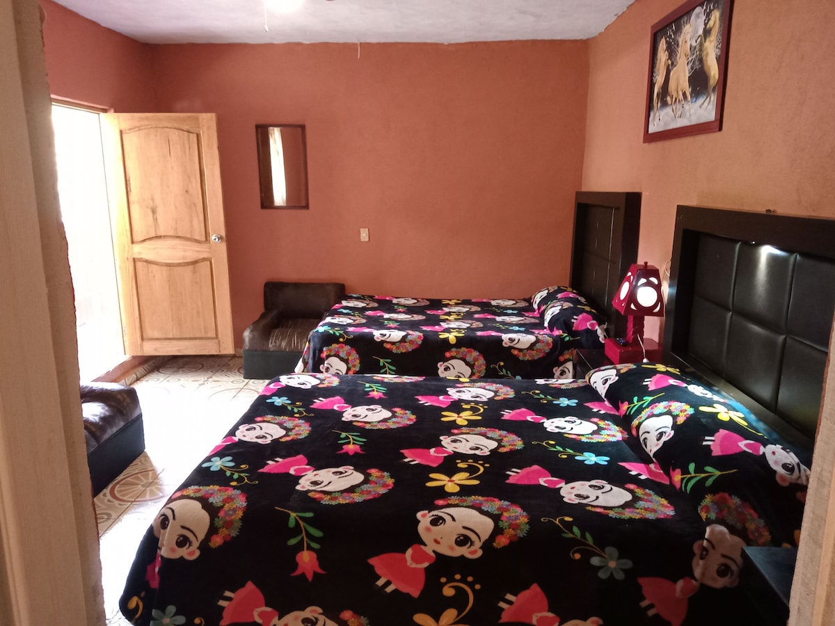 A neatly arranged sleeping area features two beds adorned with colorful blankets depicting floral designs. The warm-toned walls create a cozy ambiance, complemented by a large mirror and a wooden door leading outside. A small seating area is placed against one wall.