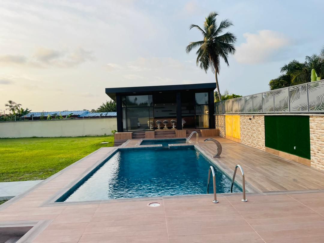 A tranquil swimming pool is framed by a wooden deck, with steps leading into the water. A modern structure with large glass windows overlooks the pool area, while green grass and palm trees enhance the outdoor setting.