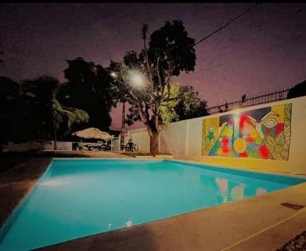 An elegantly illuminated swimming pool glows in turquoise under evening light, surrounded by a paved area. A vibrant mural featuring geometric shapes and colors provides a striking backdrop, complemented by nearby trees and outdoor seating for gathering.