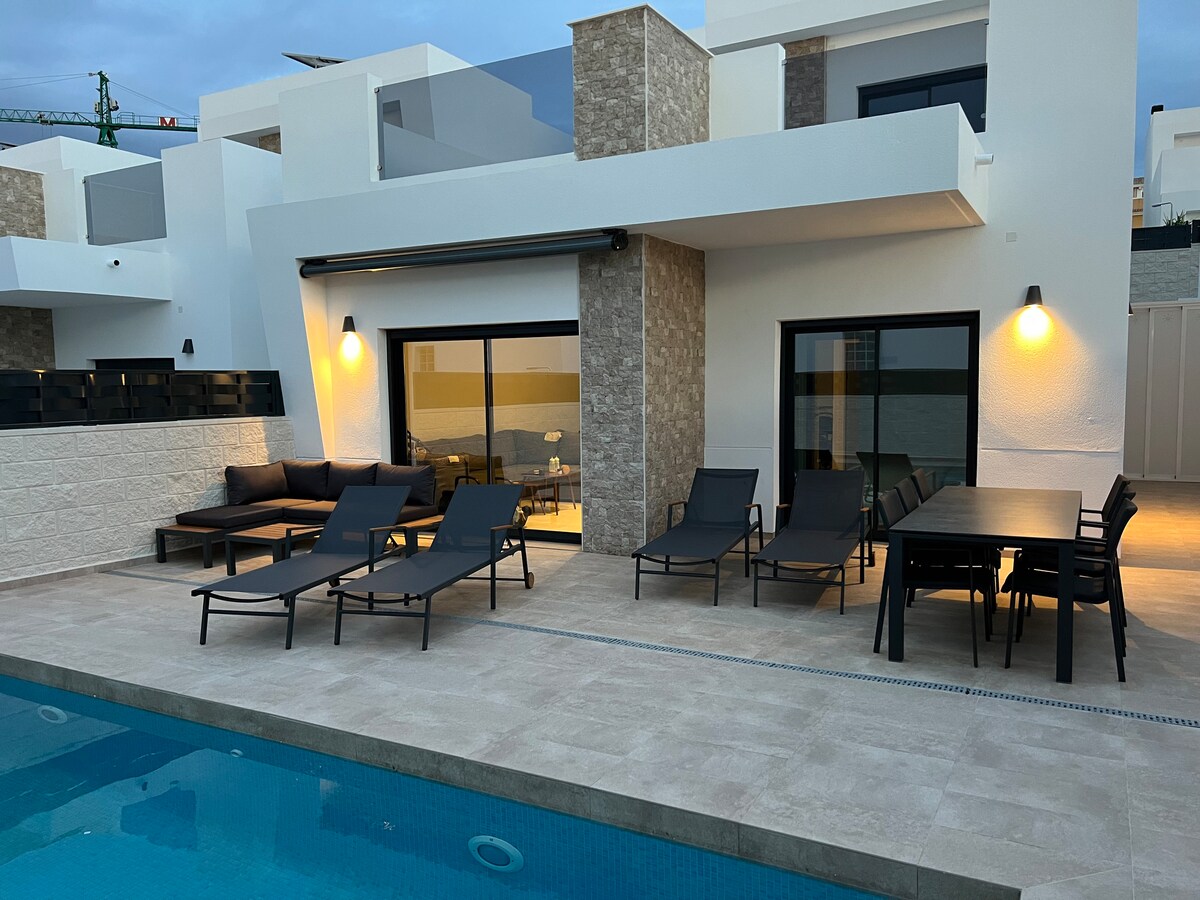 An outdoor terrace features lounge chairs arranged along a private pool. A dining table with chairs sits nearby, illuminated by wall-mounted lights. Large glass doors provide a view into the interior living space, blending comfort and relaxation in a contemporary setting.