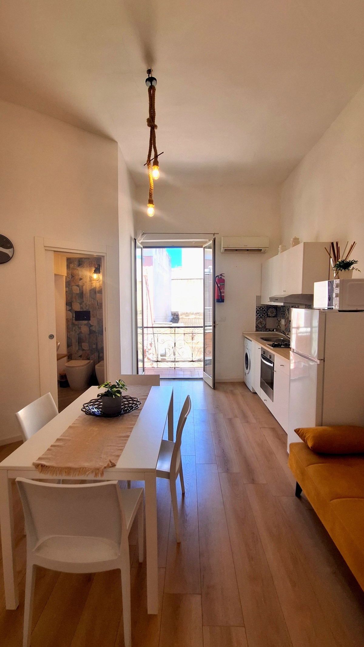 A well-lit dining area features a white table with four chairs, accompanied by a cozy yellow couch. Large glass doors open to a small balcony, allowing natural light to fill the space. A modern kitchen is visible, offering essential appliances and a decorative plant on the table.