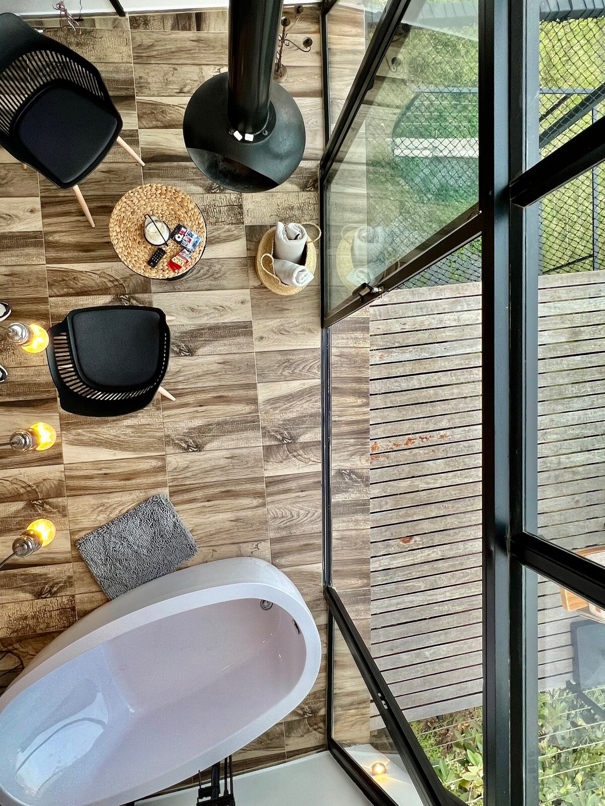 An overhead view captures a relaxing space featuring a freestanding bathtub positioned near large glass windows. Black chairs are arranged for comfort, accompanied by a woven basket and scattered towels. The warm glow from candles creates a serene ambiance, enhancing the inviting atmosphere.
