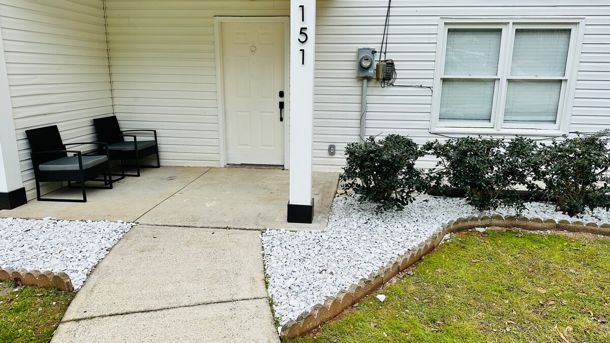 A welcoming entrance features a light-colored front door framed by decorative shrubbery. Two black chairs are positioned on the concrete porch, creating a comfortable seating area. A landscaped path, bordered by gravel and stones, leads towards the grassy area and building entrance.