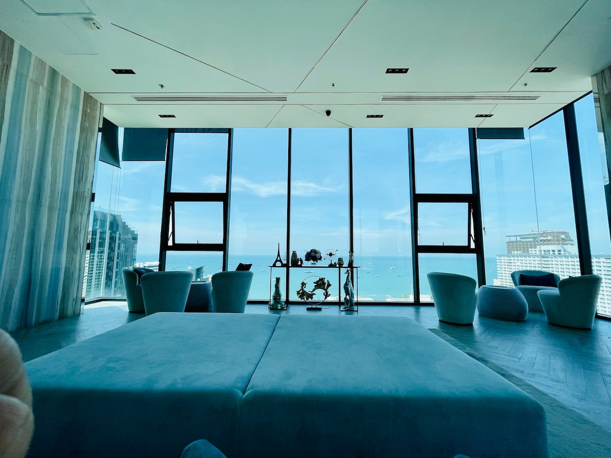 A spacious living area is highlighted by large floor-to-ceiling windows, providing a panoramic view of the ocean. Soft, circular seating arrangements complement a low, square coffee table, creating an open and bright atmosphere. Light tones in the decor enhance the sense of space and tranquility.