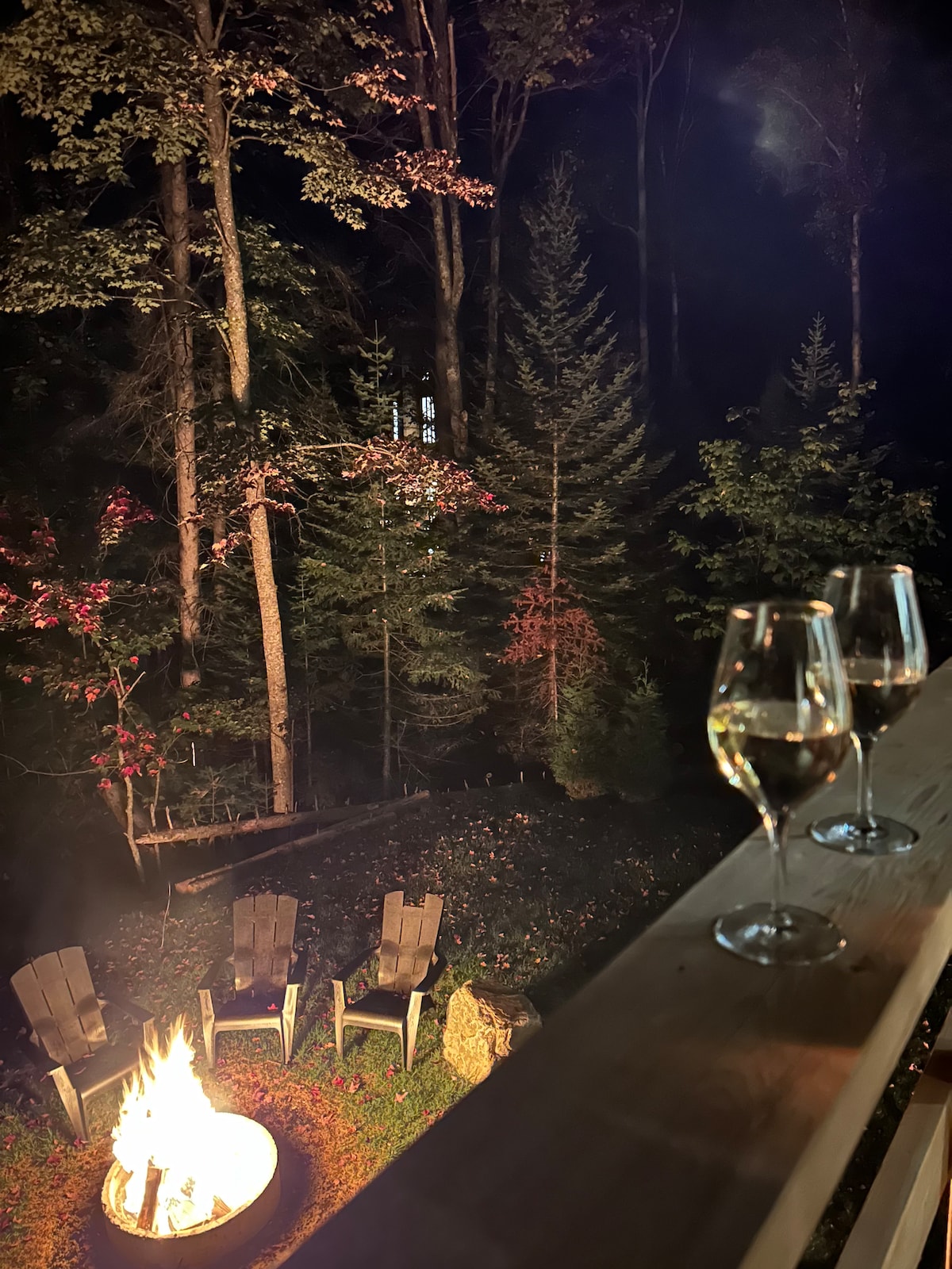 A fire pit surrounded by four adirondack chairs is shown in a wooded area, illuminated by the flames. Two glasses of white wine rest on a railing, suggesting a cozy atmosphere amidst tall trees with hints of autumn foliage.