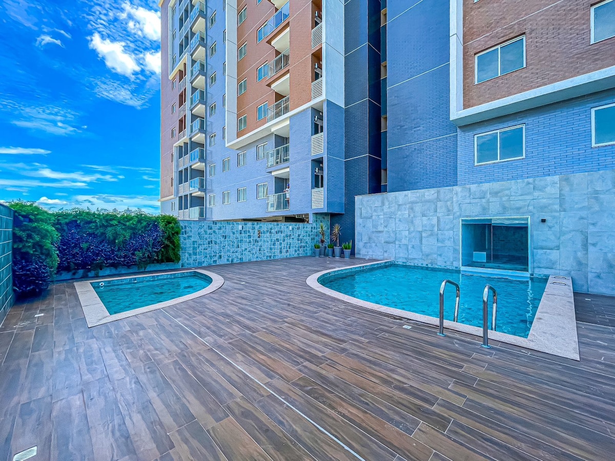 An outdoor leisure area is displayed, featuring two swimming pools surrounded by a wooden deck. Lush greenery is seen along the perimeter, while the building's modern facade looms in the background, complemented by clear blue skies.