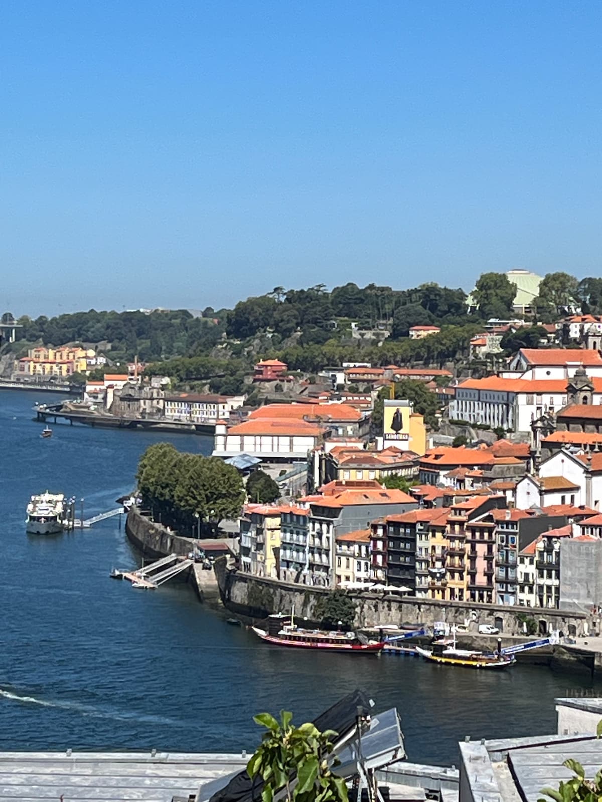The image captures a scenic view of a river winding through a town, lined with colorful buildings featuring red roofs. Boats can be seen on the water, while lush greenery adorns the hills in the background, enhancing the setting against a clear blue sky.