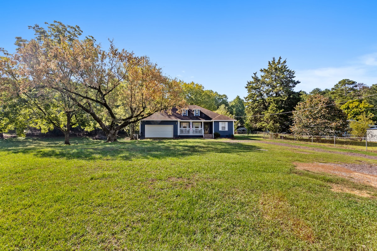 A welcoming single-story home is set within a spacious grassy area, framed by mature trees. The exterior features a covered porch with large windows, allowing natural light to illuminate the interior. A gravel drive leads up to the home, enhancing its inviting curb appeal.
