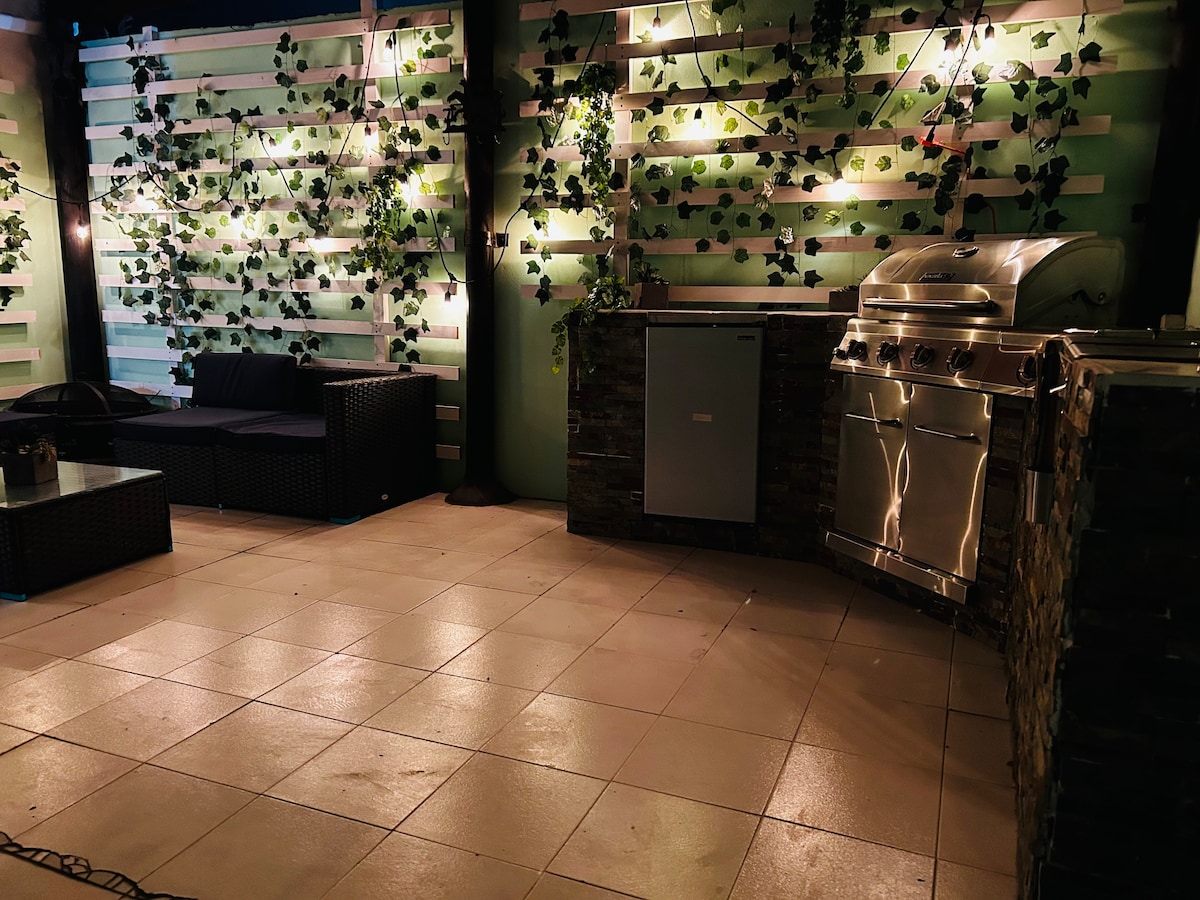 A private patio area is equipped with a modern grill and a mini refrigerator. Comfortable seating set against walls adorned with greenery creates an inviting space for relaxation. Soft lighting highlights the features, enhancing the atmosphere for outdoor gatherings.
