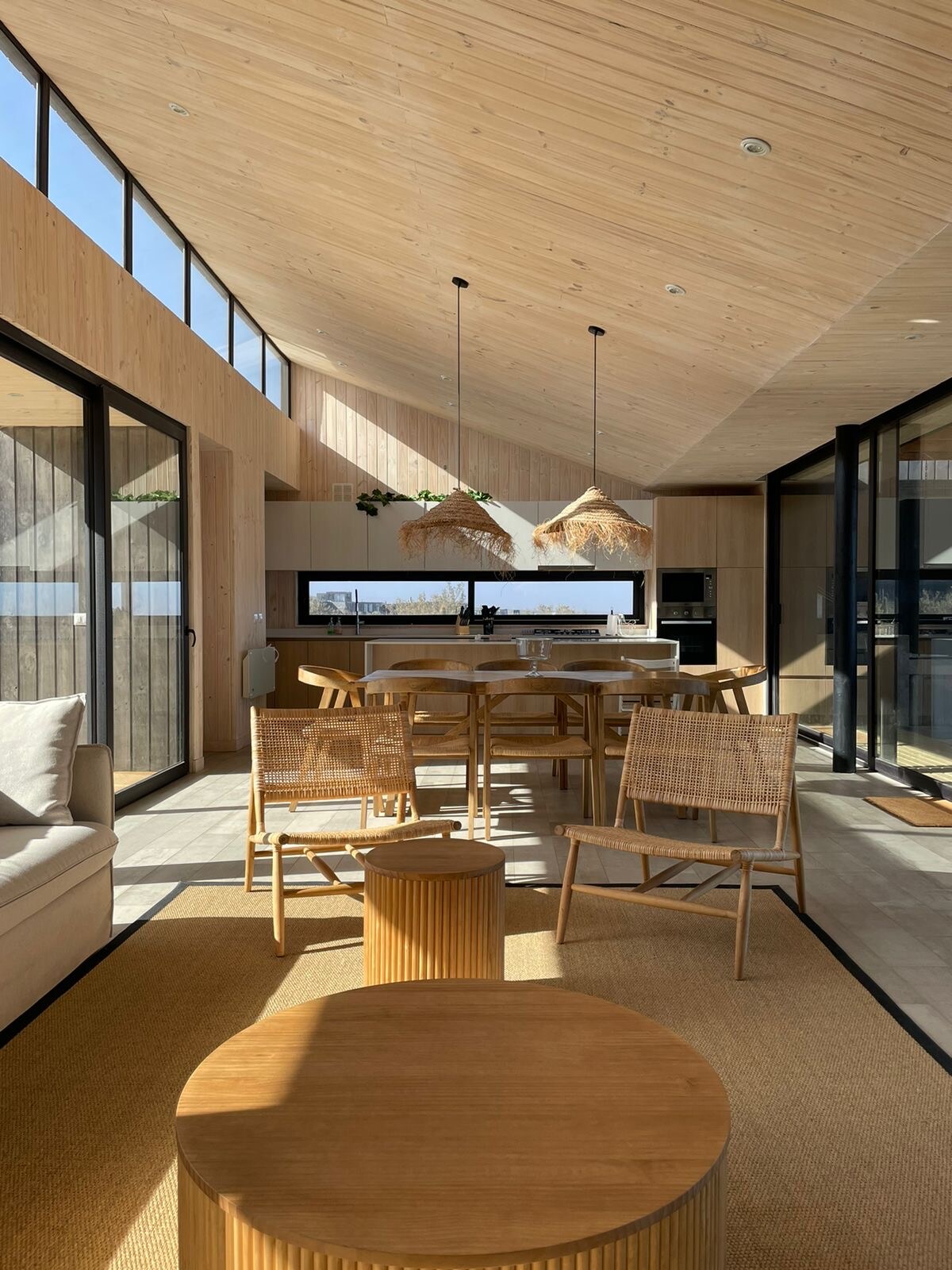 An open dining area features a wooden table surrounded by woven chairs. Large windows allow natural light to fill the space, highlighting the high wooden ceiling. Ventilation is provided through sliding glass doors, connecting the interior with the outdoor environment.