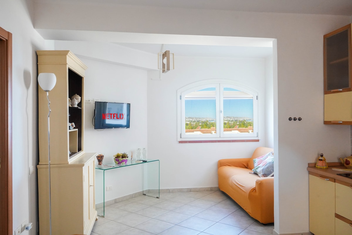 The living area features a comfortable orange sofa and a glass coffee table. A smart TV is mounted on the wall, displaying a streaming service. Large windows provide a panoramic view of the sea, filling the space with natural light.