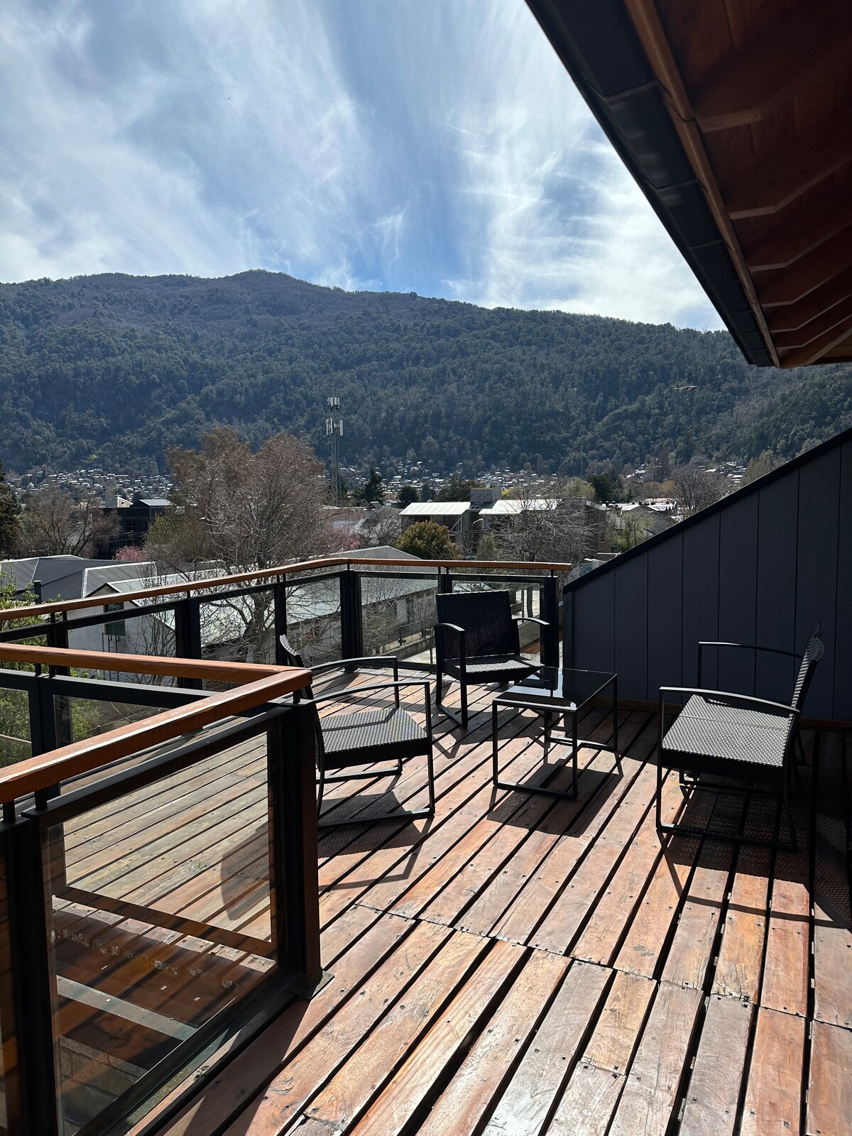 An outdoor terrace is presented with wooden decking and metal-framed chairs, offering panoramic views of the surrounding mountains and trees. Large windows provide natural light, while the sloping roof adds an architectural detail to the space.