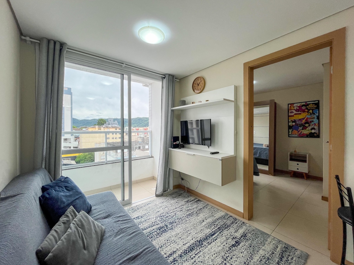 A light-filled living area features a comfortable gray sofa adorned with blue cushions. A mounted television is positioned on a white shelf. Large windows offer a view of the city, while soft gray curtains add a touch of warmth to the space.
