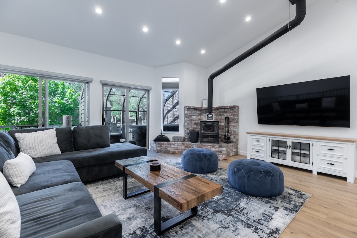 A spacious living area features a large sectional sofa in dark grey, complemented by two round blue poufs. A wooden coffee table sits in the center, with a brick fireplace and a 75-inch television mounted on the wall. Natural light pours in through the large windows.