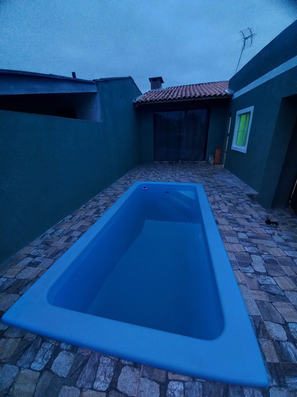 A rectangular swimming pool is situated within a paved outdoor area, surrounded by green walls. The pool features shallow steps leading into the clear blue water. A textured stone floor completes the space, with a muted sky visible above.