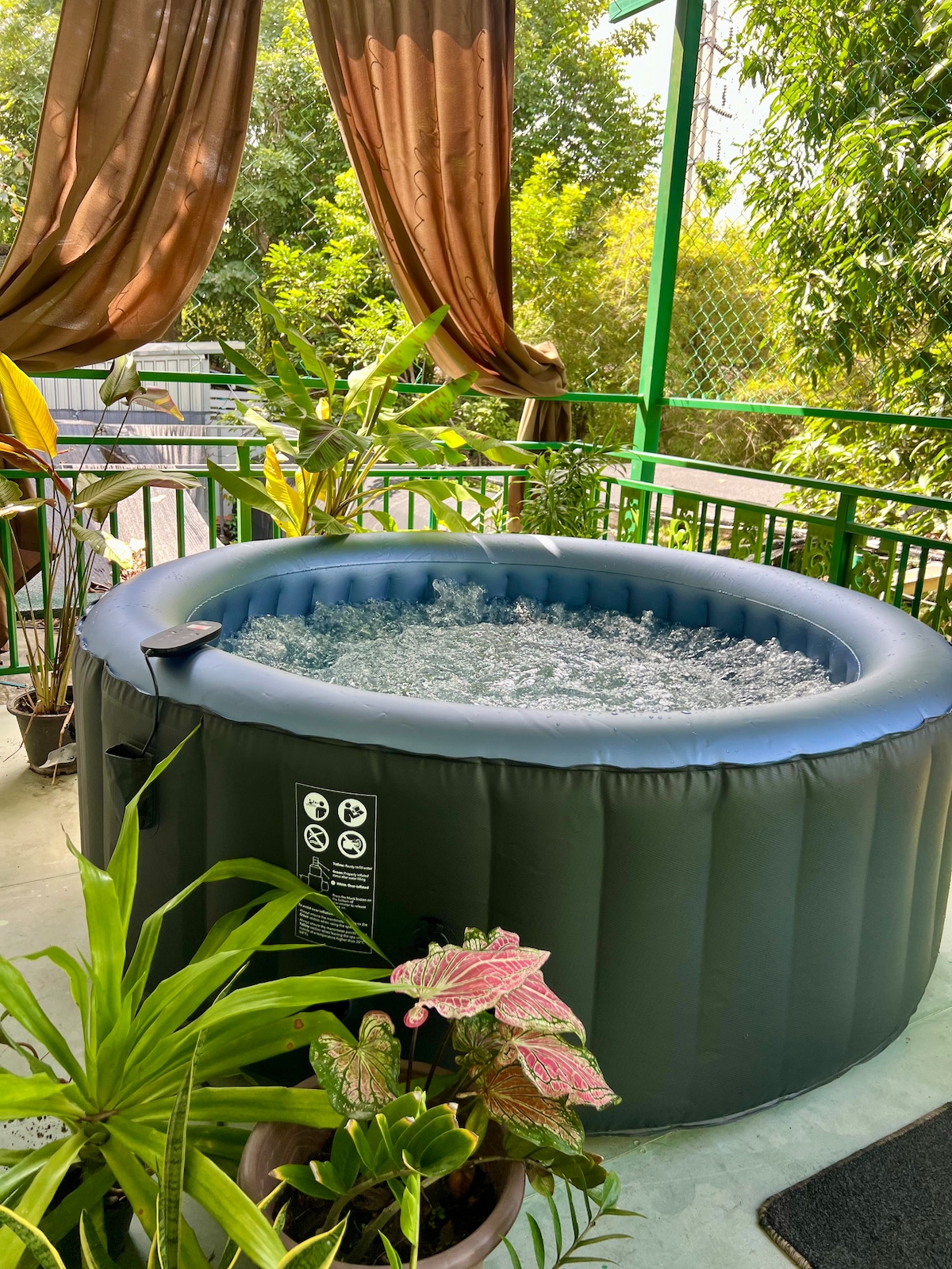 An inflatable hot tub is positioned on a patio surrounded by lush greenery. Decorative plants in various pots add a touch of nature, while curtains gently drape from the overhead structure, creating a cozy retreat space.