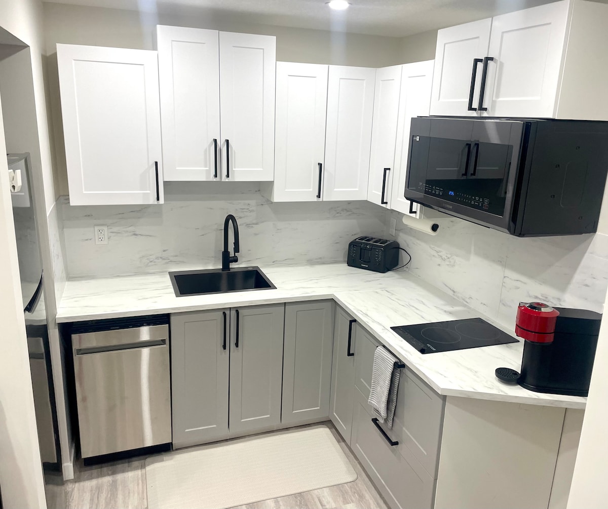 The kitchen features modern cabinetry in white and gray, complemented by a marble countertop. Stainless steel appliances include a refrigerator, dishwasher, and a microwave. A cooktop is integrated into the workspace, which is well-lit, enhancing visibility for meal preparation.
