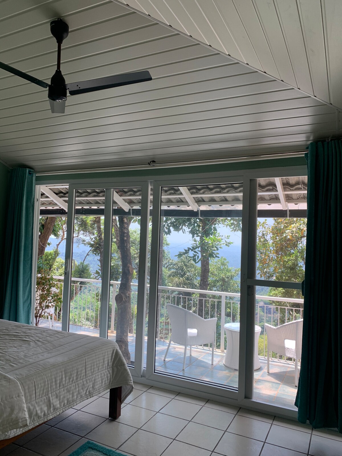 An inviting bedroom features a spacious bed covered with a white linen. Large glass doors provide access to a balcony, framed by light teal curtains. Outside, outdoor seating is visible against a backdrop of lush greenery and distant hills.