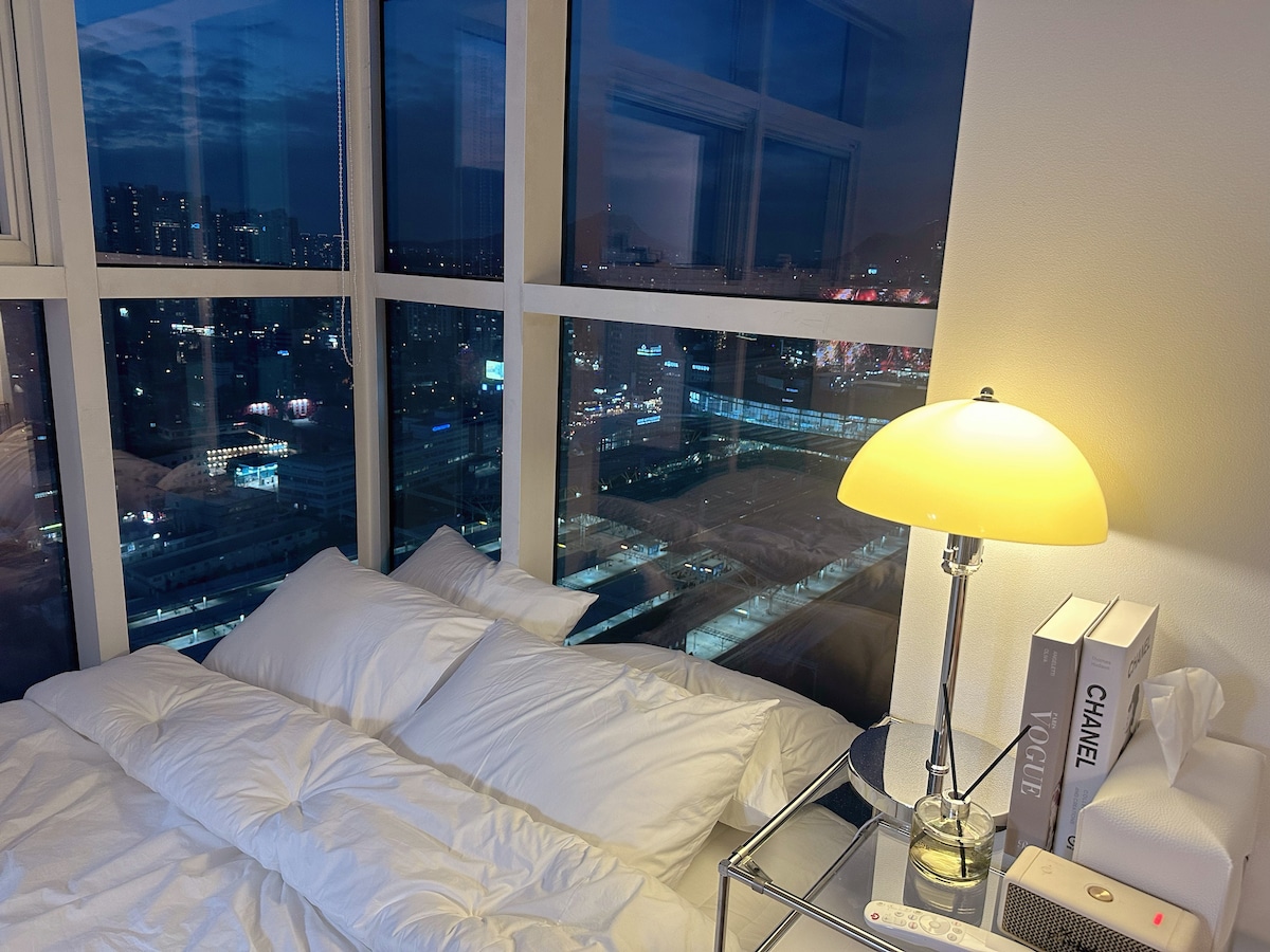 A cozy sleeping area is illuminated by a soft bedside lamp. Large windows offer a panoramic view of the city skyline at dusk, showcasing a blend of urban lights and reflections. Neatly arranged pillows and a soft bedding duvet enhance the inviting atmosphere.