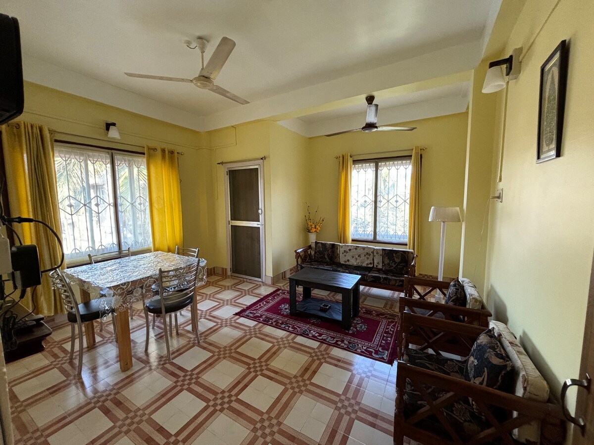 The living room features a dining table set for four, complemented by two comfortable seating arrangements. Large windows with yellow curtains allow natural light to fill the space. A decorative rug adds warmth to the tiled floor, while ceiling fans contribute to air circulation.