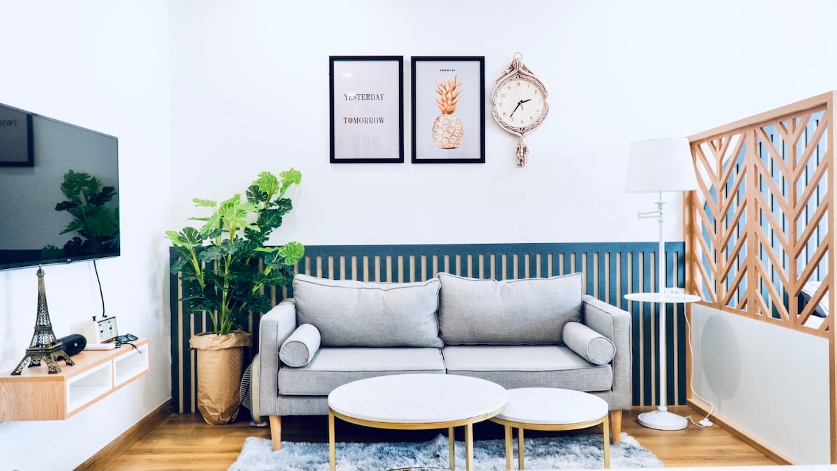 A light-filled living area features a gray sofa against a white wall adorned with framed art prints. A round coffee table occupies the center, surrounded by a potted plant and a decorative wall partition. A wall clock adds a functional touch to the space.
