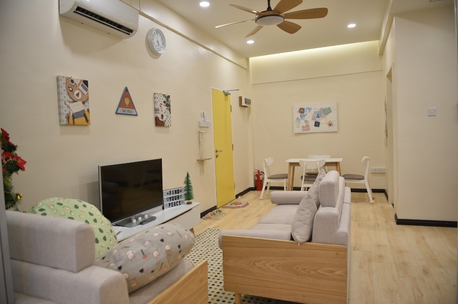 A modern living area features a light-colored sofa set with patterned cushions. A flat-screen television is positioned in front of the seating. Light wood flooring contrasts with the pale yellow walls, where artwork is displayed, and a small dining table with white chairs is visible in the background.