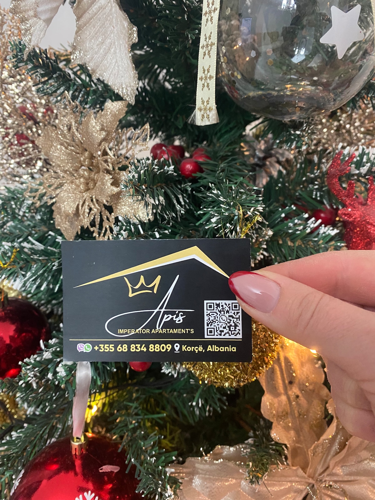 A hand is holding a business card in front of a decorated Christmas tree. The tree features gold and red ornaments, clear glass baubles, and twinkling lights, creating a festive atmosphere. The card displays contact information along with a QR code.