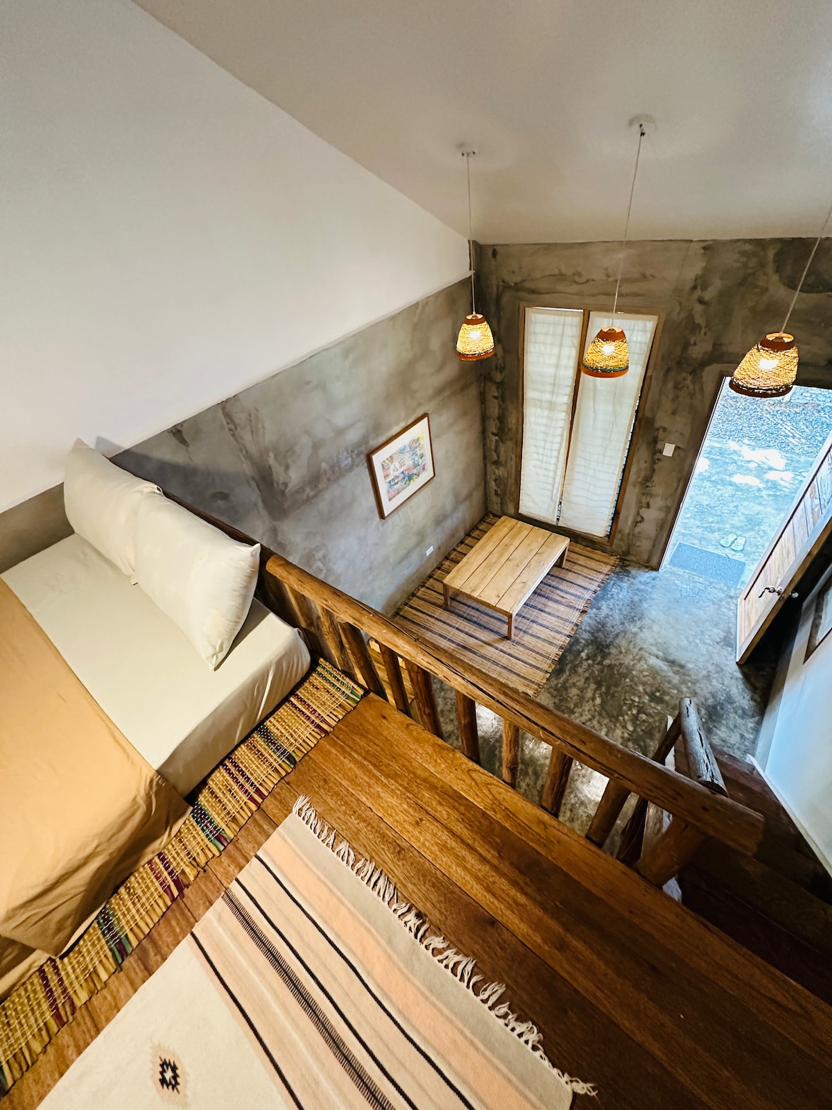 An overhead view shows a cozy sleeping area distinguished by an unmade bed with neutral linens. A wooden coffee table is positioned on a textured rug below, with textured walls and decorative lighting adding to the atmosphere. Natural light brightens the space from an open door.