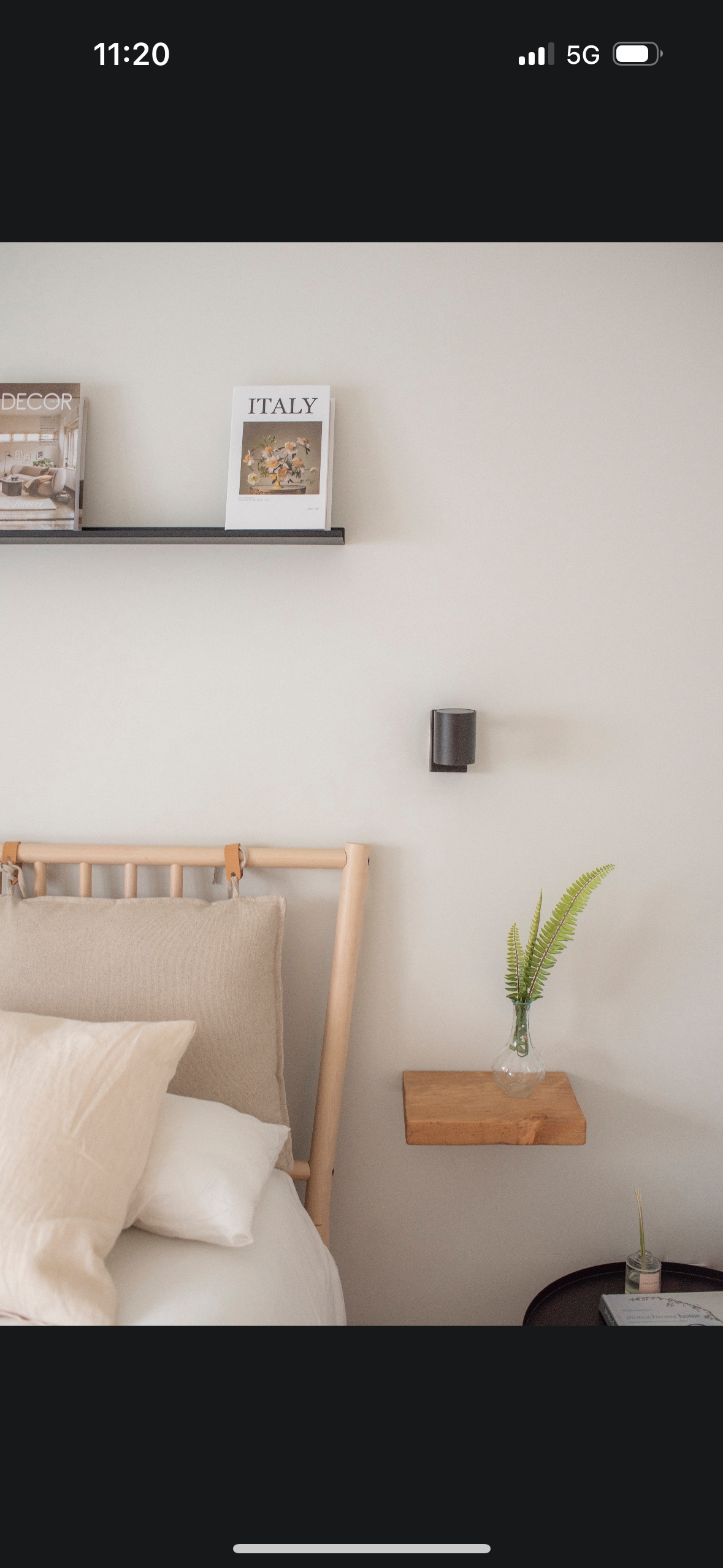 A serene bedroom setting features a light wooden bed frame with soft, neutral bedding. A small wooden shelf displays travel books above the bed, alongside a mounted wall lamp. A compact side table holds a decorative plant, enhancing the tranquil atmosphere.