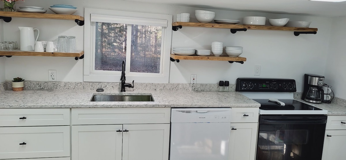 A modern kitchen is displayed, featuring a spacious countertop with a marble finish. Open wooden shelves hold white dishware, while a sleek black faucet is positioned above the stainless steel sink. A range and coffee maker are also visible, enhancing the functional space.