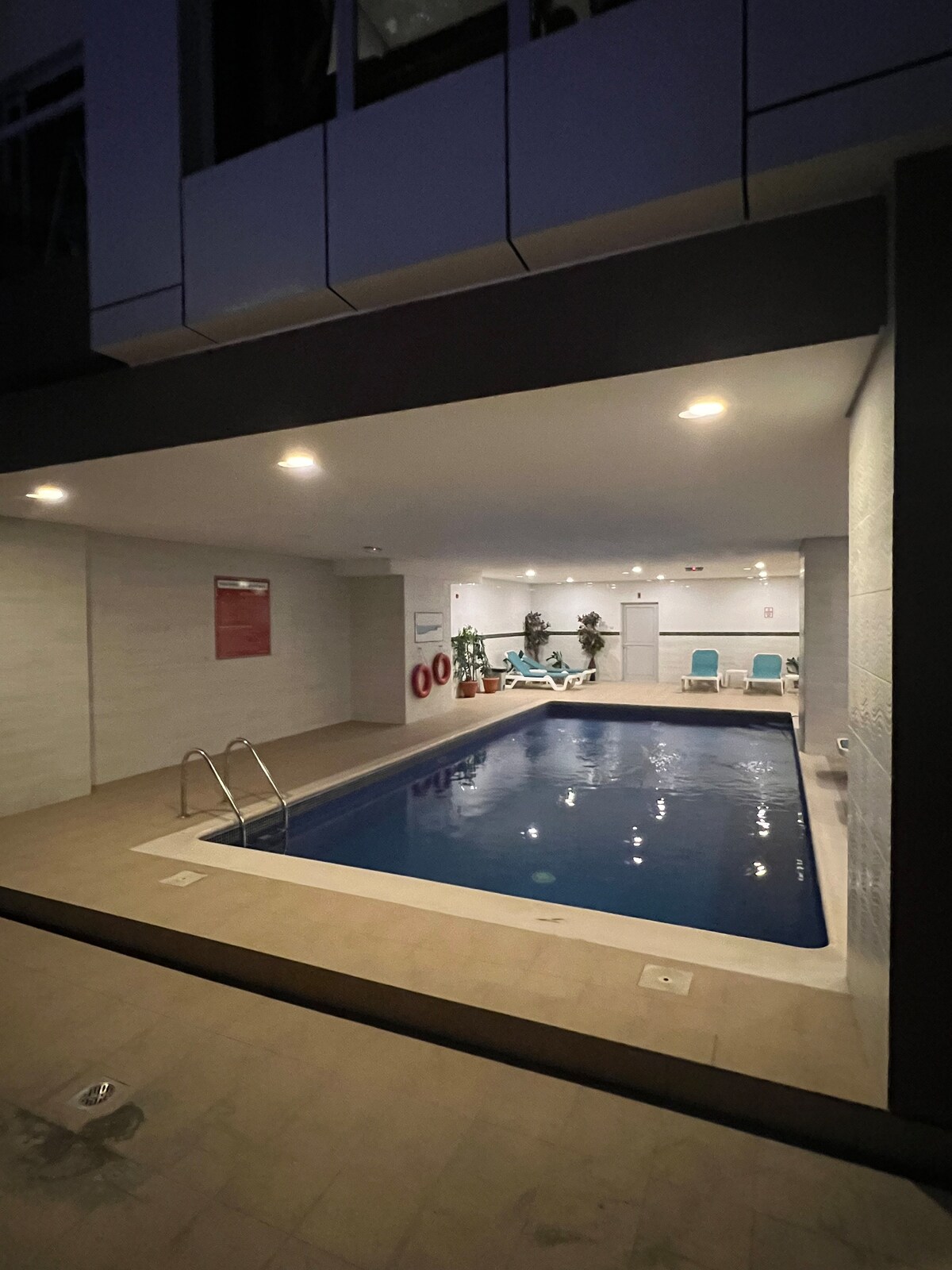 An indoor pool area is presented, featuring clear blue water and bright overhead lighting. Lounge chairs are arranged along one side, and plants add a touch of greenery. Safety equipment is visibly positioned on the walls, ensuring a secure environment.