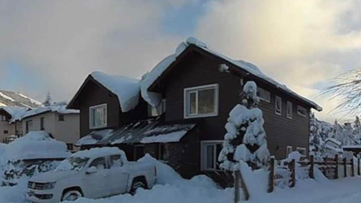 A two-story house is partially obscured by a thick layer of snow, with snow-covered trees surrounding the entrance. The roof is laden with snow. A white vehicle is parked in front, and the sky shows hints of blue amidst a clouded backdrop.