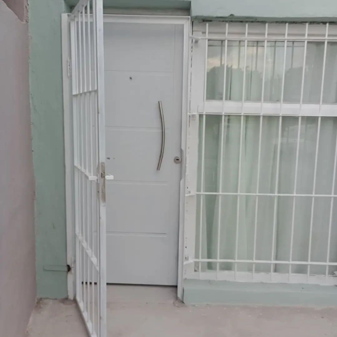 A modern entrance features a white door with a sleek handle and protective metal grilles. Large windows beside the door allow natural light to enter, enhancing the welcoming aspect of the exterior.