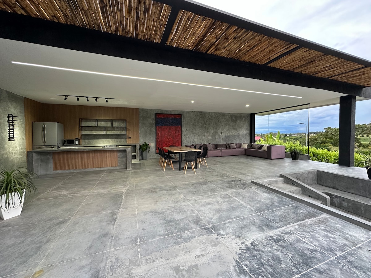 An open-concept living area features polished concrete flooring and a modern kitchen with sleek cabinetry. A spacious dining table accommodates multiple guests. Comfortable seating is arranged in a corner, with large glass doors offering views of the surrounding greenery.