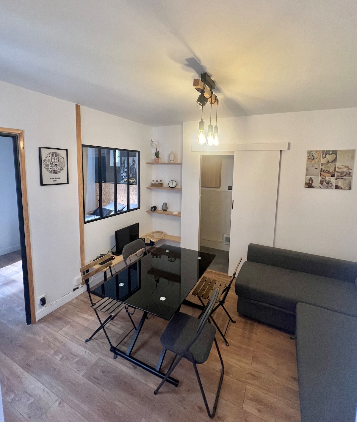 An open living area is shown with a stylish glass dining table surrounded by black chairs. The corner features a grey sectional sofa. Shelves hold decorative items, while warm light fixtures provide illumination. A glimpse of the adjoining space is visible through a window.