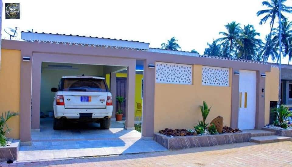 A modern exterior view showcases a single-story home with a car parked in the covered garage. The building features a mix of light yellow and white walls, with decorative cutouts adding visual interest. Lush greenery and small plants are positioned around the entrance.